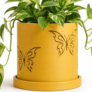 May include: A yellow cylindrical planter with a matching saucer, featuring cut-out butterfly designs. The planter holds a green plant with variegated leaves. The plant's vines cascade over the sides, creating a decorative and natural aesthetic.