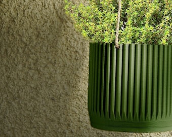 May include: A 5 inch matte green hanging planter with a ribbed design. The planter is hanging from a rope and has a green plant inside.