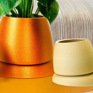May include: Three decorative planters in shades of yellow and gold sit on a matching table. The planters are holding a lush green plant with large, patterned leaves. The background includes a gray sofa.