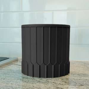 May include: A black ceramic utensil holder with a fluted design. The holder is sitting on a countertop.