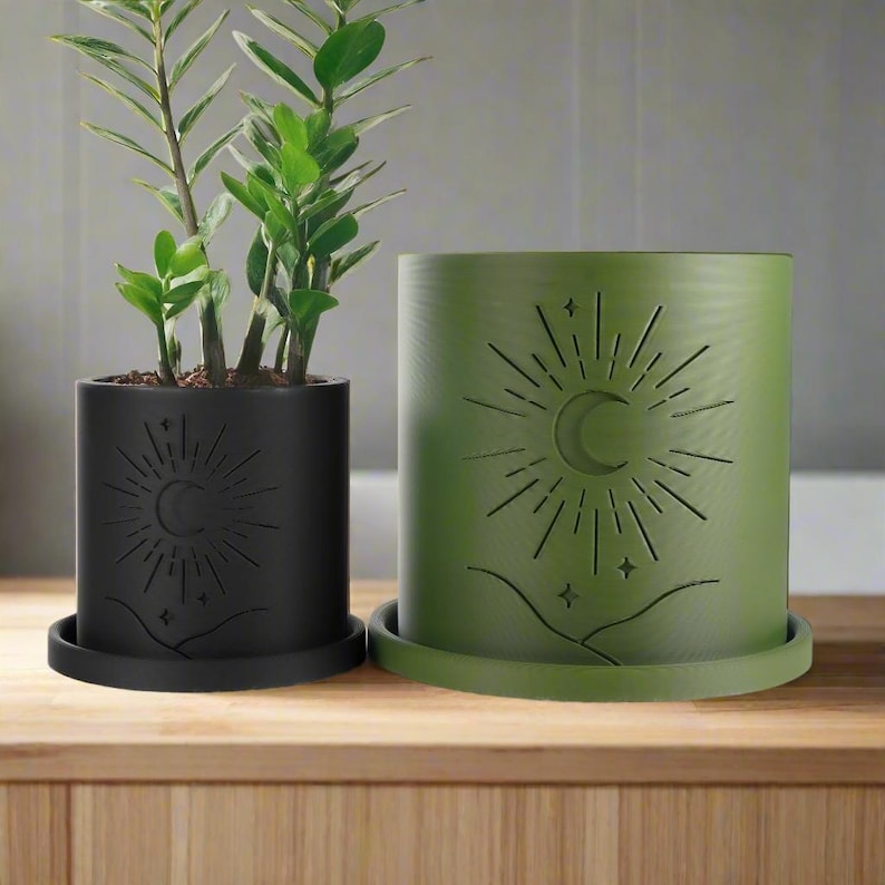 Op de afbeelding: Twee potplanten met een maan- en zonontwerp. De kleinere pot is zwart en de grotere pot is groen.