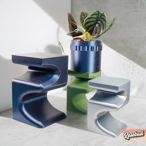 May include: Three abstract, modern, sculptural stools in blue, green, and silver. The stools are shaped like the letter 'S' and are made of a smooth, glossy material. A potted plant sits on top of the green stool.