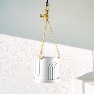 May include: White ceramic hanging planter with a rope hanger and an abstract design of arches. The planter has a white saucer.