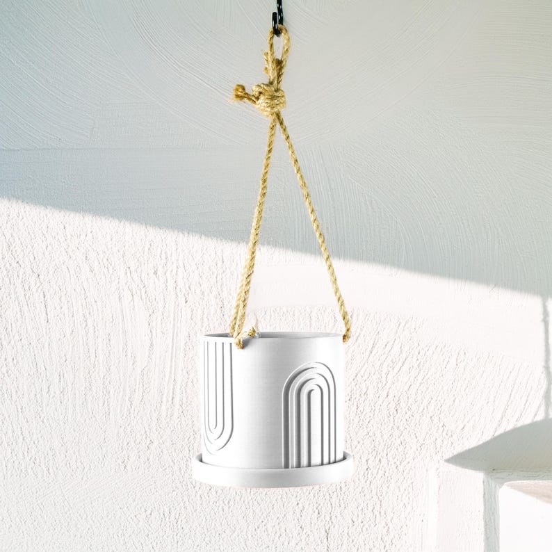 May include: White ceramic hanging planter with a rope hanger and an abstract design of arches. The planter has a white saucer.