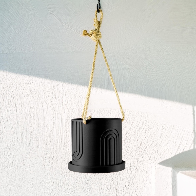 May include: Black hanging planter with a rope and a geometric design. The planter is made of ceramic and has a matte finish.