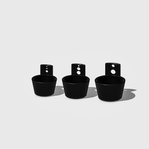 May include: Three black wall-mounted cup holders. Each holder has a round cup shape and a rectangular back plate with two holes for hanging.