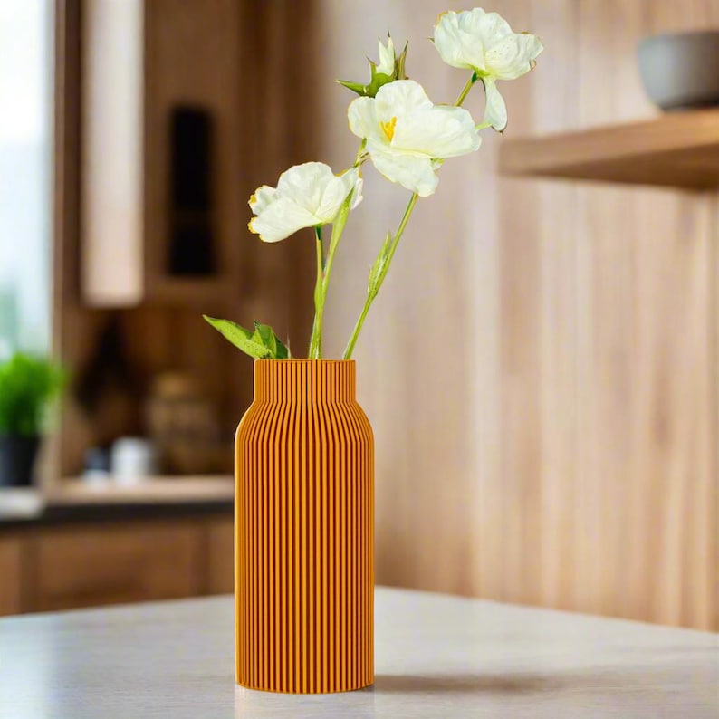 May include: A tall, orange vase with vertical grooves, holding a bouquet of white flowers. The vase is on a white table.