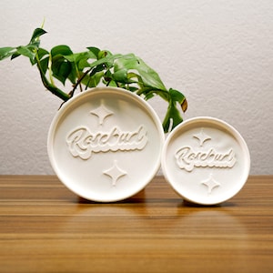 May include: Two white round containers with the word "Rosebud" embossed on them. The larger container is in the background and the smaller container is in the foreground.