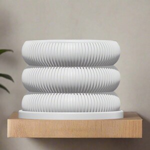 May include: A white ceramic planter with a ribbed design. The planter is stacked in three tiers and sits on a wooden shelf.