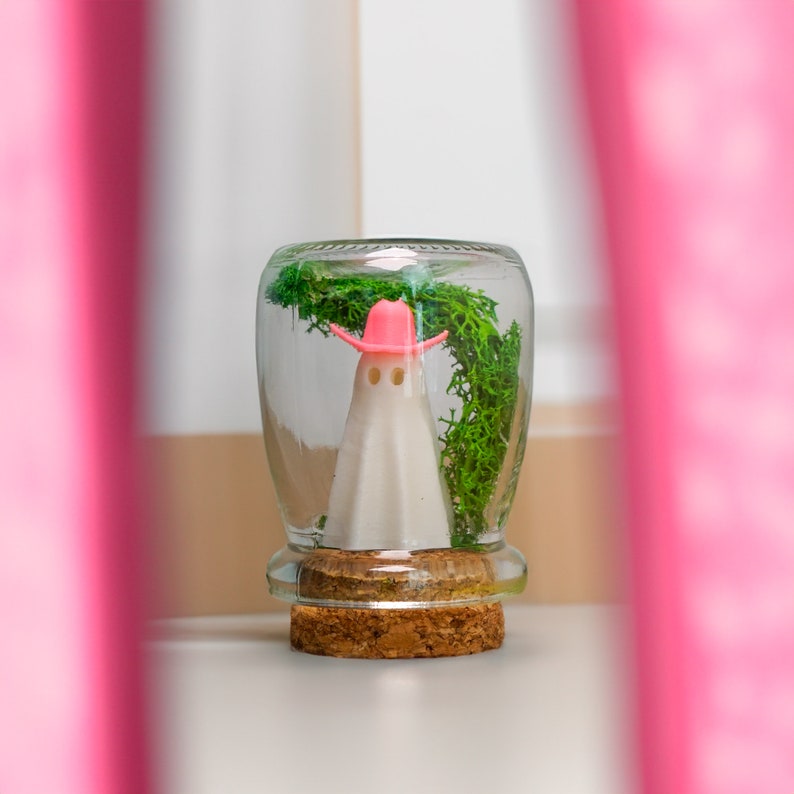 May include: A clear glass jar with a cork lid contains a white ghost figurine wearing a pink cowboy hat. Green moss is visible around the ghost.