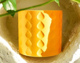 May include: A cylindrical, bright yellow planter with a repeating teardrop pattern. The planter is set in a textured, off-white alcove, with green leaves in the background. The planter is designed for indoor or outdoor use.