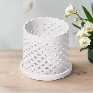Puede incluir: Maceta de cerámica blanca con un diseño de rejilla decorativo. La maceta tiene una base ancha y plana y una forma cilíndrica.