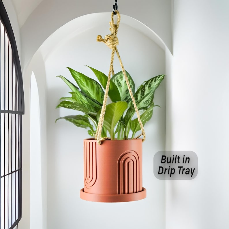 May include: A terracotta hanging planter with a built-in drip tray and a geometric design. The planter is hanging from a rope and has a green plant inside.