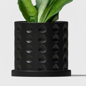 May include: A black planter with a textured raindrop pattern. The planter has a wide base and a cylindrical shape. A green plant is growing inside the planter.