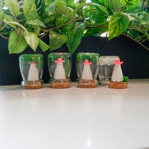 May include: Four glass jars with cork lids, each containing a small white ghost wearing a pink cowboy hat and green moss.