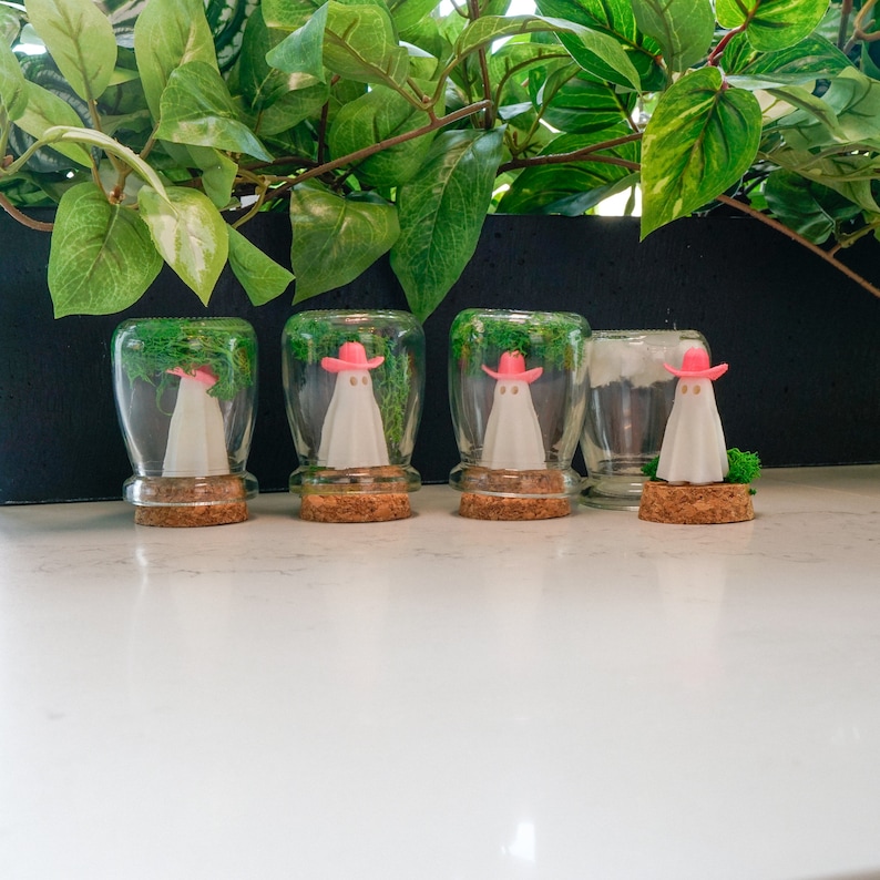 May include: Four glass jars with cork lids, each containing a small white ghost wearing a pink cowboy hat and green moss.