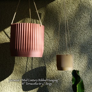 May include: Two hanging planters, one terracotta and one beige, with a ribbed design. The terracotta planter is 8 inches in diameter and the beige planter is 4 inches in diameter. The planters are hanging from a light brown rope.
