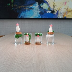 May include: Four glass jars with cork lids, each containing a small, white figurine wearing a pink cowboy hat and green moss. The jars are arranged in a row on a light wood surface.