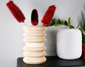 May include: A white ceramic vase with a wavy design, holding three red dried flower stems. The vase is sitting on a dark brown wooden surface.