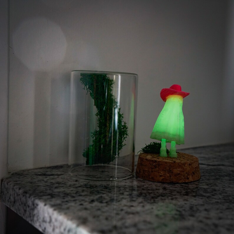 May include: A green glowing figurine wearing a red hat stands on a cork base. The figurine is in front of a glass jar with green moss inside.