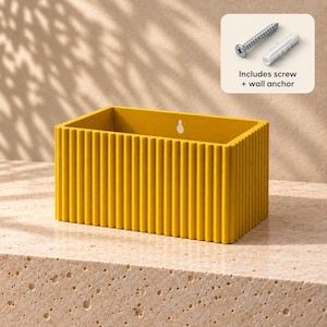 Puede incluir: Una maceta rectangular de pared en color amarillo mostaza con una textura acanalada vertical en el exterior. El panel trasero incluye una ranura en forma de cerradura para el montaje. Incluye un tornillo metálico y un taco de pared blanco.