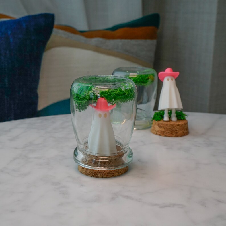 May include: Two glass jars with cork lids, each containing a small white ghost figurine with a pink hat. The jars are filled with green moss and are sitting on a white marble surface.