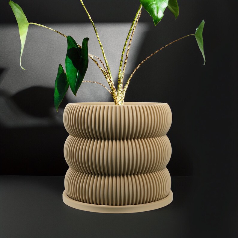 May include: A beige ceramic planter with a unique, layered, ribbed design. The planter is filled with a green plant with large leaves.