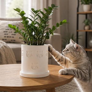 May include: A white ceramic planter with a cat face design, holding a green plant. A gray tabby cat reaches toward the plant. The planter sits on a round wooden table in a well-lit room. Home decor.