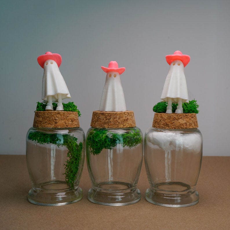 May include: Three glass jars with cork lids, each containing a small white ghost figurine wearing a pink cowboy hat and green moss.
