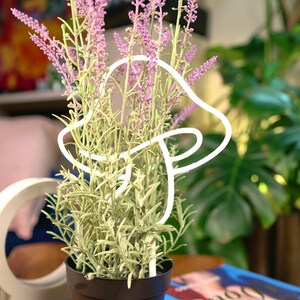 Peut inclure: Une sculpture lumineuse en forme de champignon en n&eacute;on blanc avec une plante de lavande dans un pot noir. La plante a des fleurs violettes et des feuilles vertes.