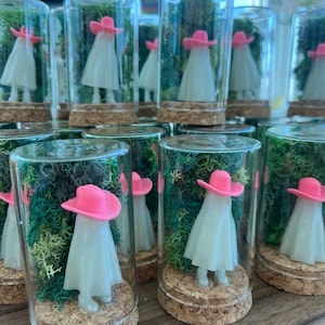 May include: A collection of glass bottles with cork tops, each containing a small white figurine wearing a pink hat. The figurines are surrounded by green moss.
