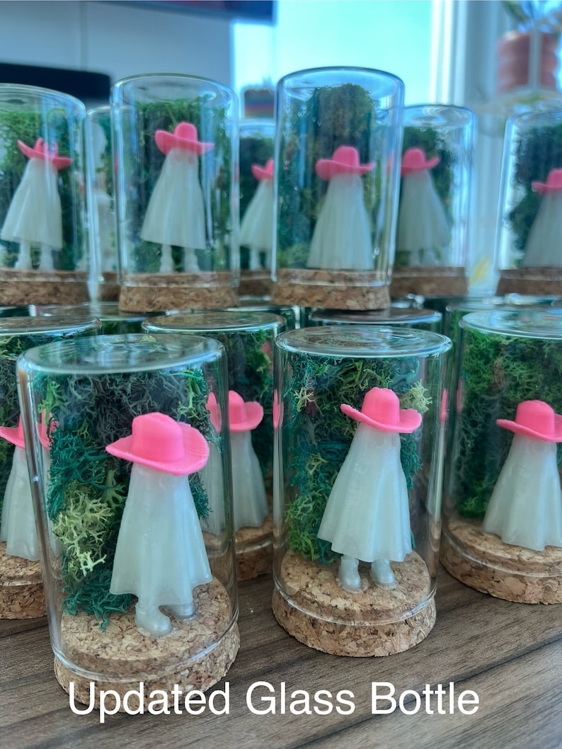 May include: A collection of glass bottles with cork tops, each containing a small white figurine wearing a pink hat. The figurines are surrounded by green moss.