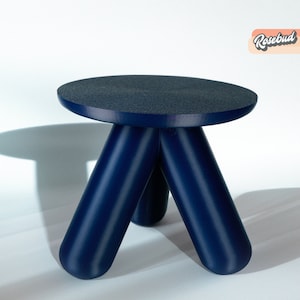 Könnte beinhalten: Ein blauer, dreibbeiniger Hocker mit einer runden, strukturierten Oberseite. Der Hocker ist aus einem glatten, glänzenden Material gefertigt.