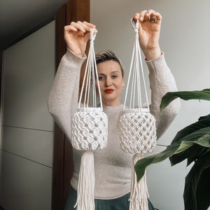 Könnte beinhalten: Eine Frau hält zwei Makramee-Pflanzenhänger. Die Hänger sind aus weißem Baumwollschnur und haben eine zylindrische Form. Die Hänger hängen von der Decke.