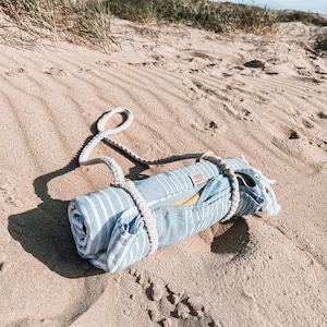 Könnte beinhalten: Ein blaues und weiß gestreiftes Strandtuch mit einem weißen Seilgriff. Das Handtuch ist aufgerollt und hat eine kleine Holzperle am Griff. Das Handtuch liegt auf einem Sandstrand.