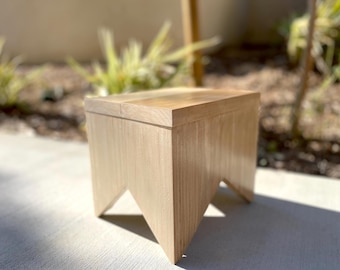 May include: A light-colored wooden side table with a rectangular top and three angled legs. The table is made of light-colored wood and has a natural finish. The table measures approximately 18 inches in height.