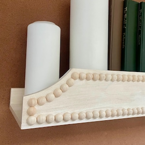 Puede incluir: Una estantería de madera clara con borde de cuentas, que contiene dos velas blancas y varios libros. Los libros tienen cubiertas verdes y beige. La estantería está montada en una pared marrón.