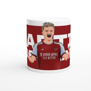 May include: White ceramic mug with a red background featuring a cartoon illustration of a soccer player in a red and blue jersey with the text "Arsenal" and "Emirates Fly Better" on the jersey. The player is celebrating with his arms raised.
