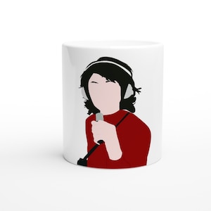 Pode incluir: Caneca de cerâmica branca com uma ilustração em preto e branco de uma pessoa usando fones de ouvido e segurando um microfone. A pessoa está vestindo uma camisa vermelha.