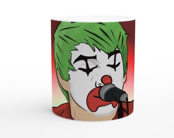 Peut inclure: Une tasse en céramique blanche avec une illustration colorée d'une personne aux cheveux verts portant un maquillage de clown et tenant un microphone. L'arrière-plan est un dégradé de rouge, orange et jaune.