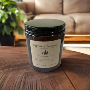 May include: A brown glass candle jar with a black lid and a paper label that reads "Coffee & Tobacco" and "EARTH MADE CO. EST. 2023". The label also includes the text "SPRINGVILLE, UTAH" and "6 OZ SOY WAX CANDLE".