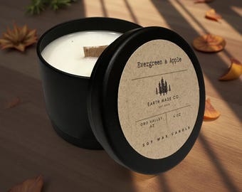 Puede incluir: Una vela de cera de soja negra en una lata con la tapa abierta. La etiqueta dice "Evergreen & Apple" y "Earth Made Co." La vela es de 113g y tiene una mecha de madera. El fondo es una superficie de madera con hojas.