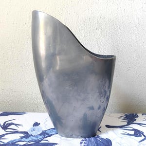 May include: A silver metal ice bucket with a distinctive curved shape. The bucket has a wide top and a narrower base. The surface reflects light, and it's placed against a white wall and a blue and white patterned fabric.