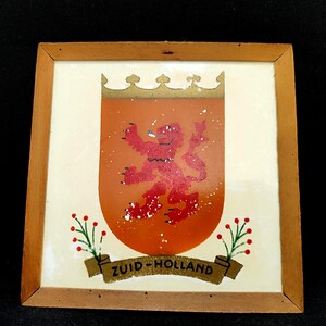 Puede incluir: Obra de arte enmarcada en madera con un escudo con un león rojo rampante sobre un fondo naranja. El escudo está coronado con una corona dorada y la pancarta dice "ZUID-HOLLAND". Ramas decorativas con bayas rojas flanquean la pancarta.