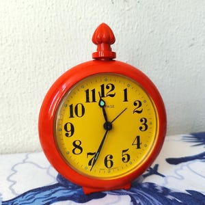 Puede incluir: Un reloj despertador rojo con una esfera amarilla y números negros. El reloj tiene un pequeño pomo rojo en la parte superior y dice "Virage" en la esfera.