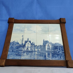Op de afbeelding: Een ingelijst tegelkunstwerk met een blauw en wit stadsgezicht met gebouwen, boten en waterreflecties. Het kunstwerk is geplaatst in een donkerbruine houten lijst met decoratieve hoekaccenten. De scène doet denken aan een historische Europese stad.