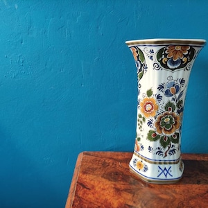 Può includere: Un vaso in ceramica bianca con un motivo floreale blu e bianco. Il vaso ha una base esagonale e una parte superiore svasata. Il vaso è appoggiato su un tavolo di legno.