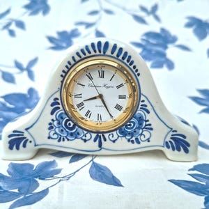 Könnte beinhalten: Eine weiße und blaue Keramik-Kaminuhr mit goldfarbenem Zifferblatt. Die Uhr hat römische Ziffern und den Markennamen "Christian Huygens". Sie ist mit blauen Blumenmustern verziert.