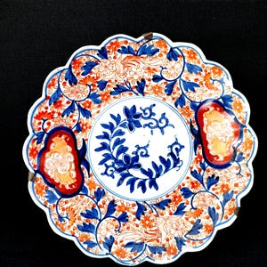 May include: A decorative ceramic plate with a scalloped edge. The plate features a floral design in blue, orange, and white. The center has a blue floral pattern, and the rim has a repeating floral pattern. Two decorative elements are on the rim.