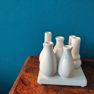 May include: A set of six white ceramic bud vases arranged in a square formation on a white base. The vases are all different shapes and sizes, and they are perfect for displaying small bouquets of flowers.
